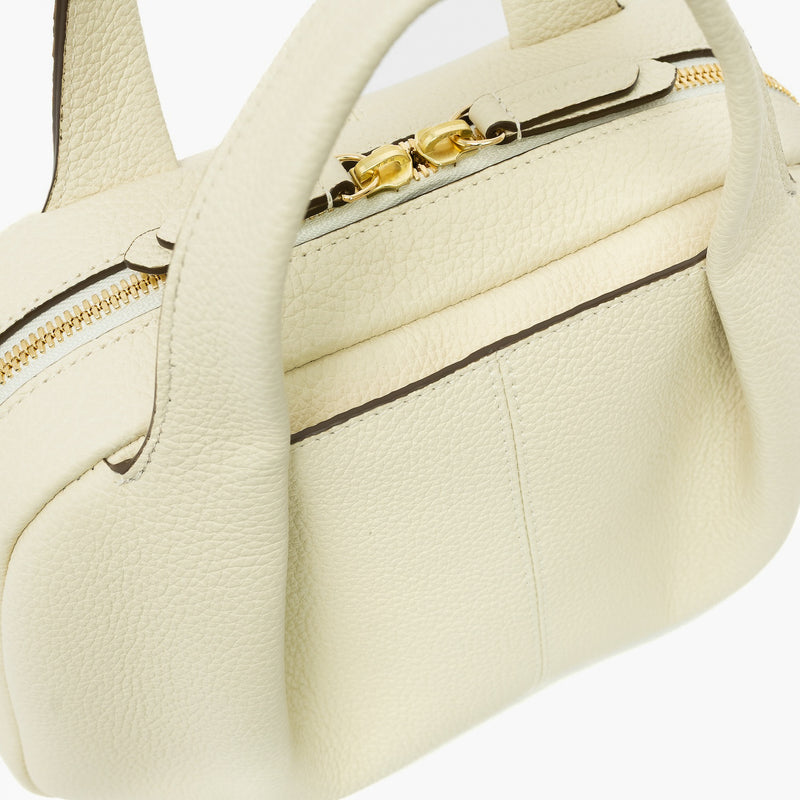 Close up of STOW London's Mini Bowling bag in Spring Moon pebbled leather featuring a pleated front panel, the zip closure and STOW London embossed pull tabs on a light grey background