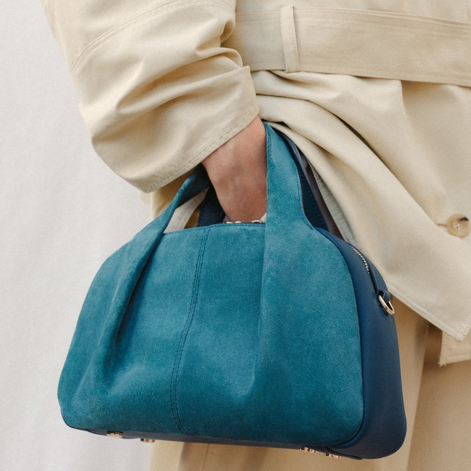 Model holding STOW London's Mini Bowling bag in Ocean Teal suede and smooth leather featuring a pleated front panel, detachable crossbody strap and gold-coated stainless steel hardware 