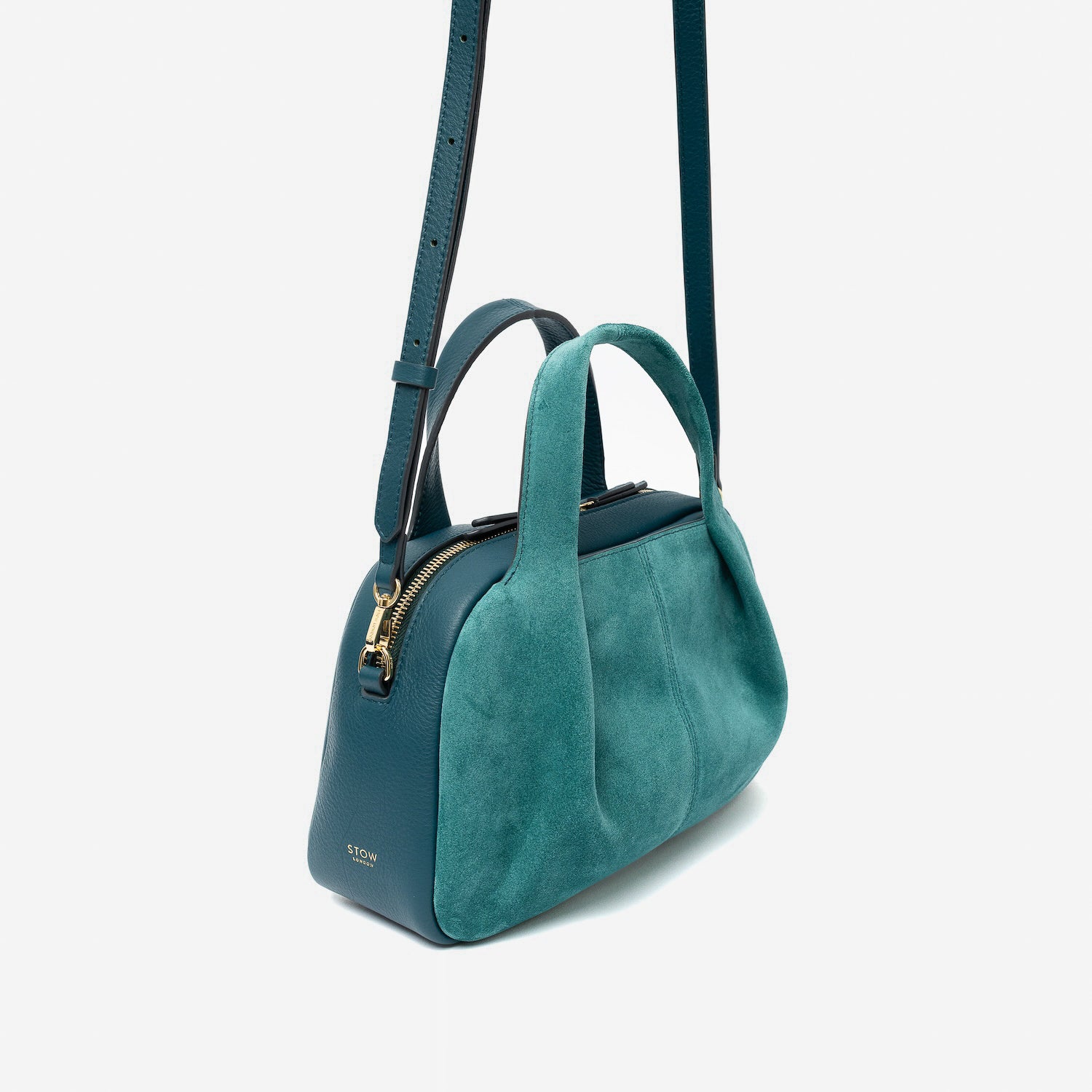 Off-axis view of STOW London's Mini Bowling bag in Ocean Teal suede and smooth leather featuring a pleated front panel, its detachable crossbody strap and gold-coated stainless steel hardware on a light grey background