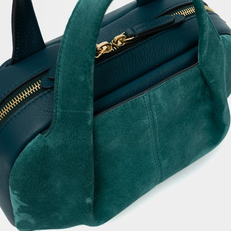 Close up of STOW London's Mini Bowling bag in Ocean Teal suede and smooth leather featuring a pleated front panel, the zip closure with STOW London embossed pull tabs on a light grey background