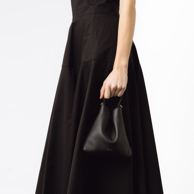 Model holding STOW London's Mini Curve in Black pebbled leather by top handle at her side on a white background 