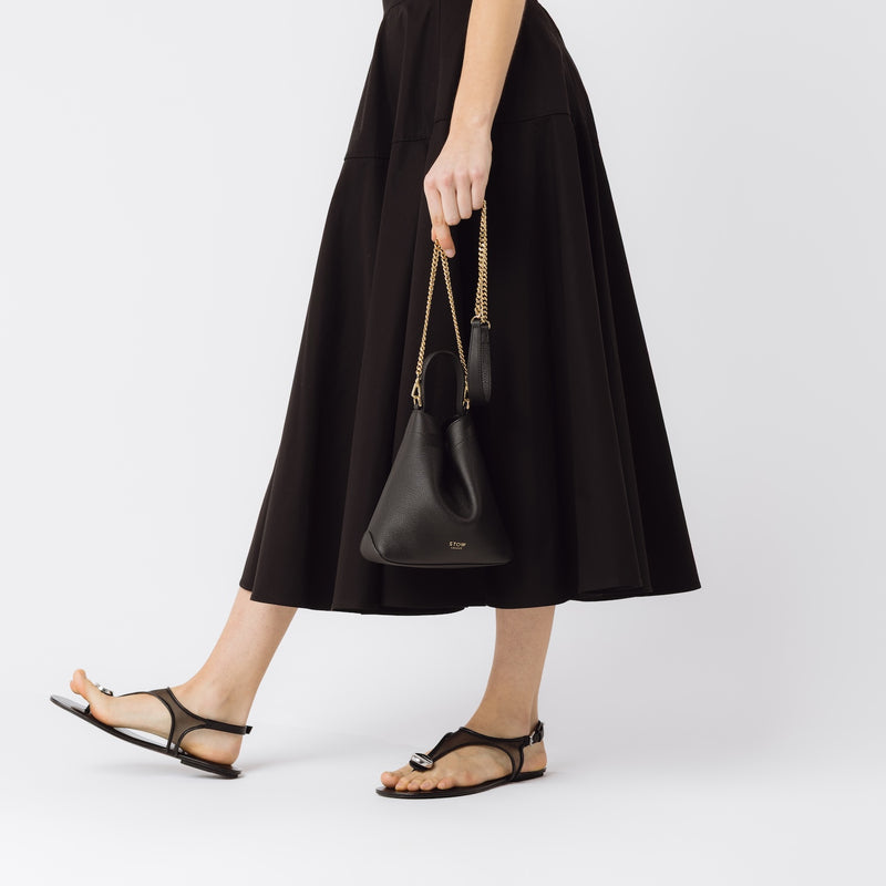 Model holding STOW London's Mini Curve in Black pebbled leather by the detachable gold chain crossbody strap at her side on a white background 