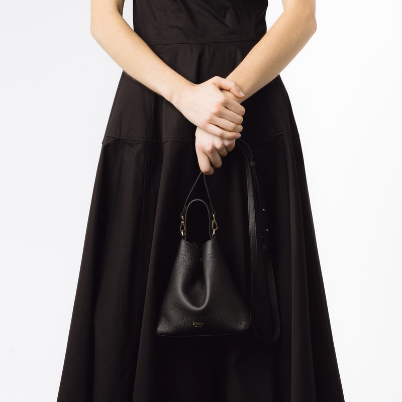 Model holding STOW London's Mini Curve in Black pebbled leather by the detachable leather crossbody strap on a white background 