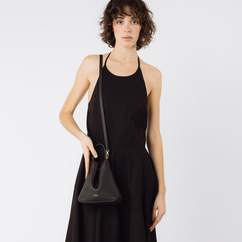 Model wearing STOW London's Mini Curve in Black pebbled leather with the detachable leather crossbody strap on her shoulder on a white background 
