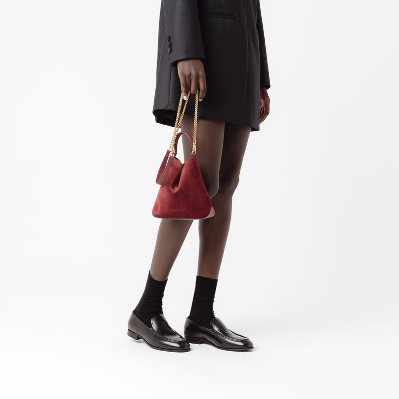 Model holding Stow London's Mini Curve in Burgundy suede leather by the detachable gold chain crossbody strap on a white background 