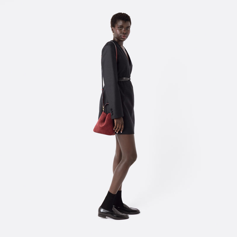 Model wearing STOW London's Mini Curve in Burgundy suede leather with the detachable leather crossbody strap on her shoulder on a white background 