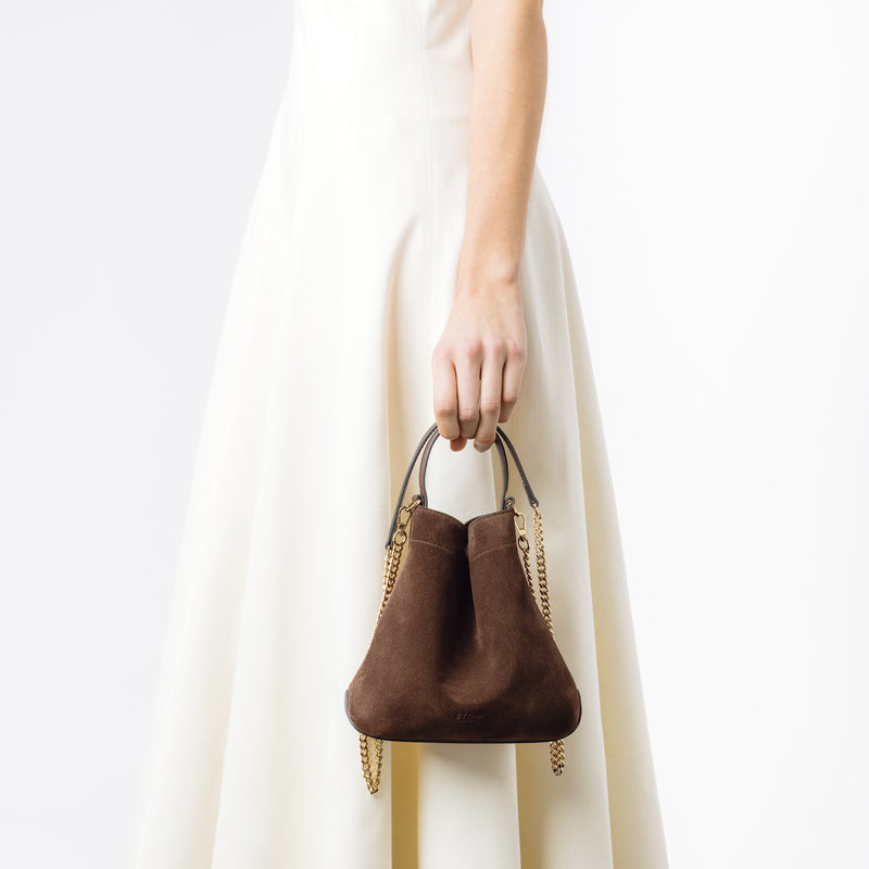 Model holding Stow London's Mini Curve in Chocolate suede leather and detachable gold chain crossbody strap at her side on a white background 