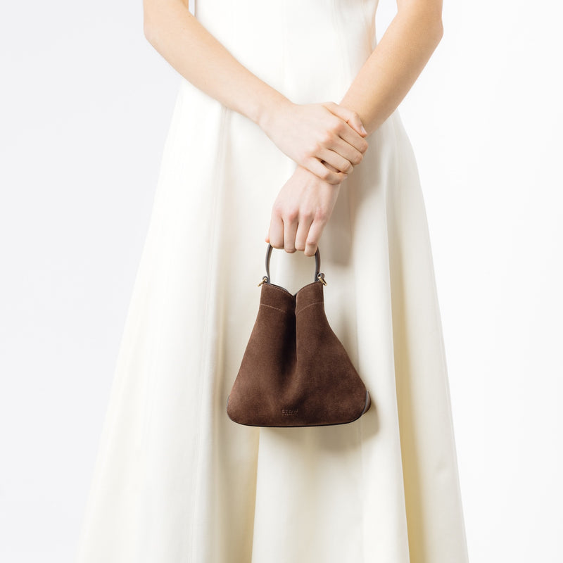 Model holding Stow London's Mini Curve in Chocolate suede leather by top handle on a white background 