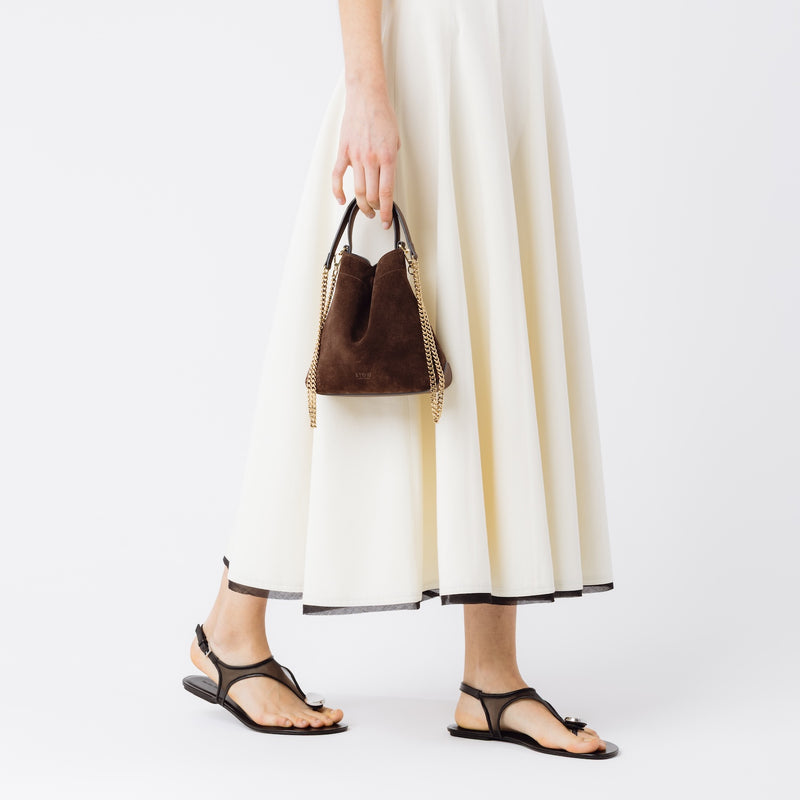 Model holding Stow London's Mini Curve in Chocolate suede leather and detachable gold chain crossbody strap at her side on a white background 