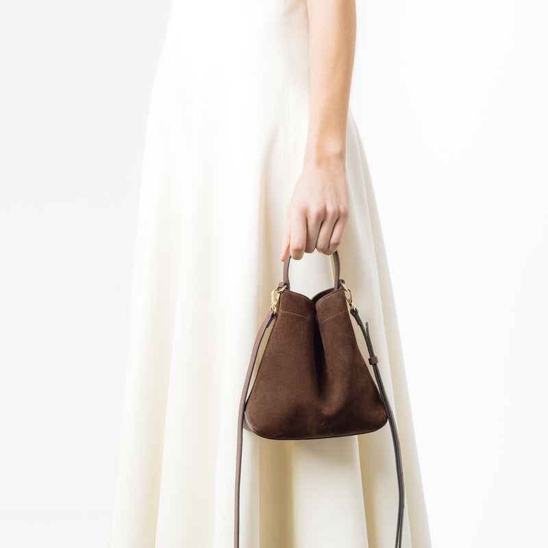 Model holding STOW London's Mini Curve in Chocolate suede leather with the detachable leather crossbody strap on a white background 