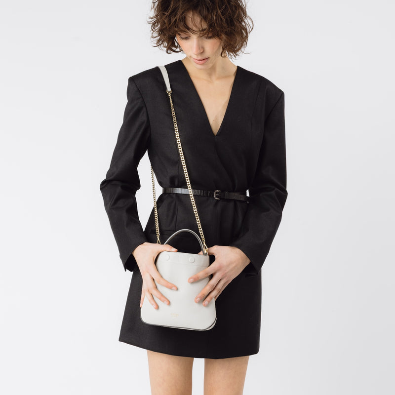 Model wearing Stow London's Mini Curve in Cloud Grey pebbled leather on her shoulder with the detachable gold chain crossbody strap on a white background 