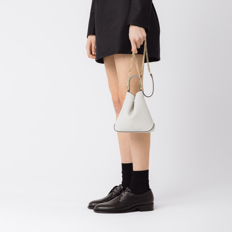 Model holding Stow London's Mini Curve in Cloud Grey pebbled leather by its detachable gold chain crossbody strap at her side on a white background 
