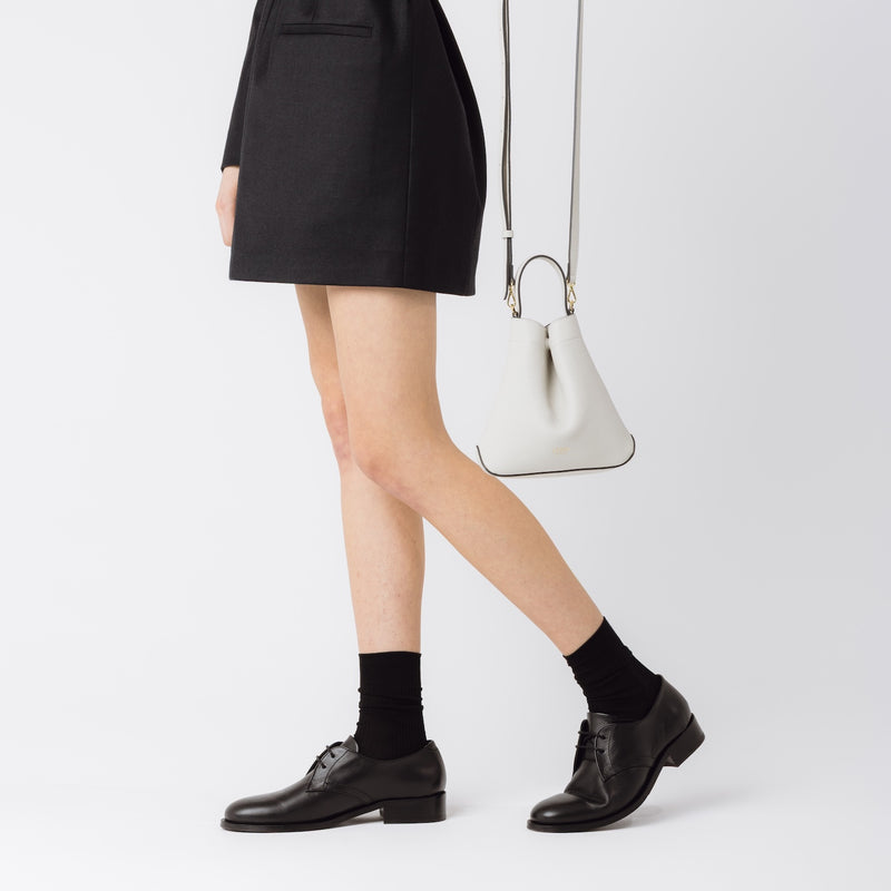 Model holding STOW London's Mini Curve in Cloud Grey pebbled leather with the detachable leather crossbody strap on a white background 