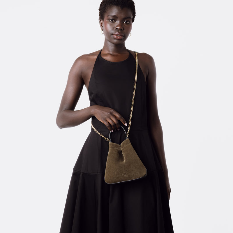 Model wearing Stow London's Mini Curve in Dark Green suede leather crossbody featuring gold-coated stainless steel hardware and chain on a light grey background 