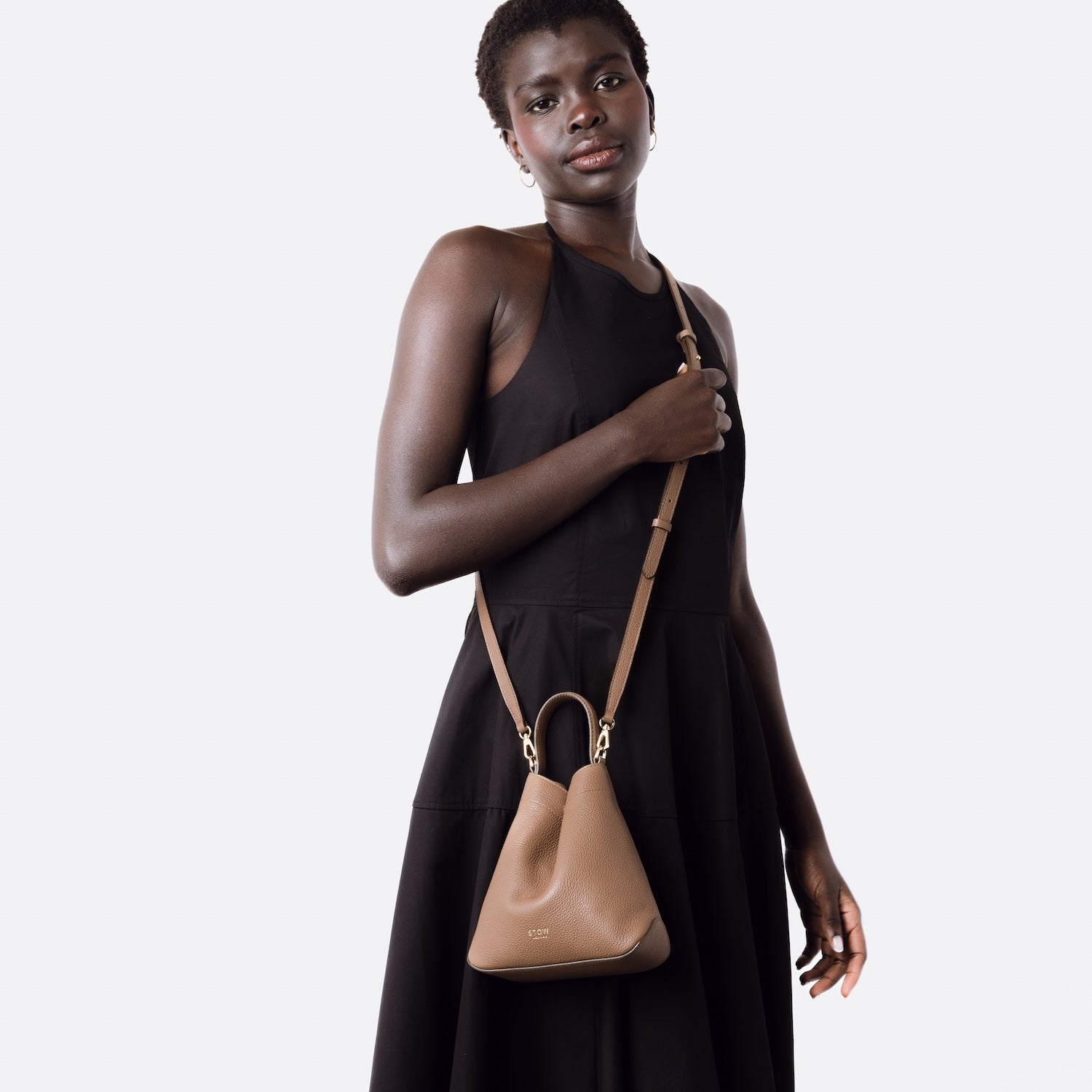 STOW London's Mini Curve in Mocha  leather worn crossbody with leather strap featuring gold-coated stainless steel hardware on a white background