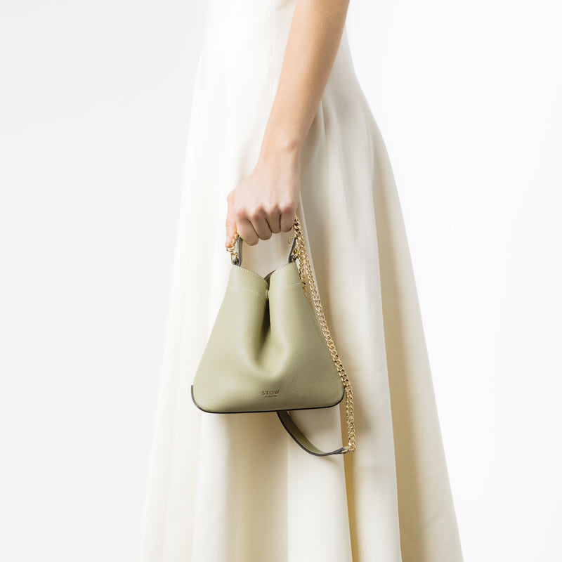 Model holding STOW London's Mini Curve in Pale Green pebbled leather and detachable gold chain crossbody strap at her side on a white background 