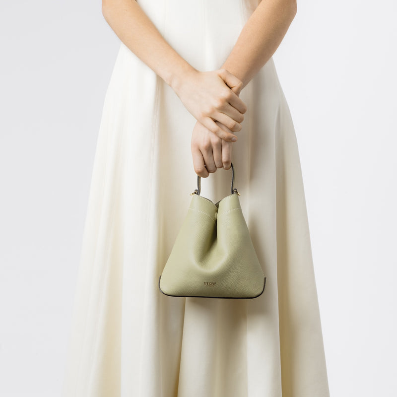 Model holding STOW London's Mini Curve in Pale Green pebbled leather by top handle on a white background 