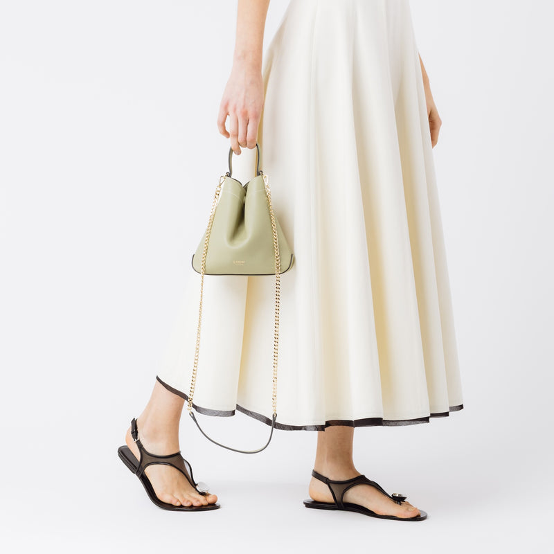 Model holding STOW London's Mini Curve in Pale Green pebbled leather and detachable gold chain crossbody strap at her side on a white background 