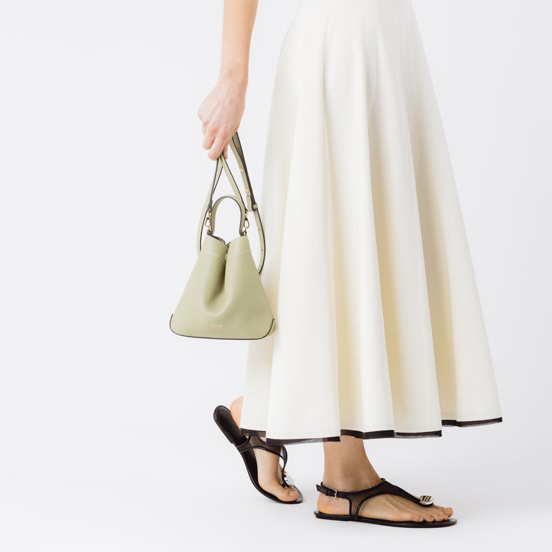 Model holding  STOW London's Mini Curve in Pale Green pebbled leather by the detachable leather crossbody strap at her side on a white background 