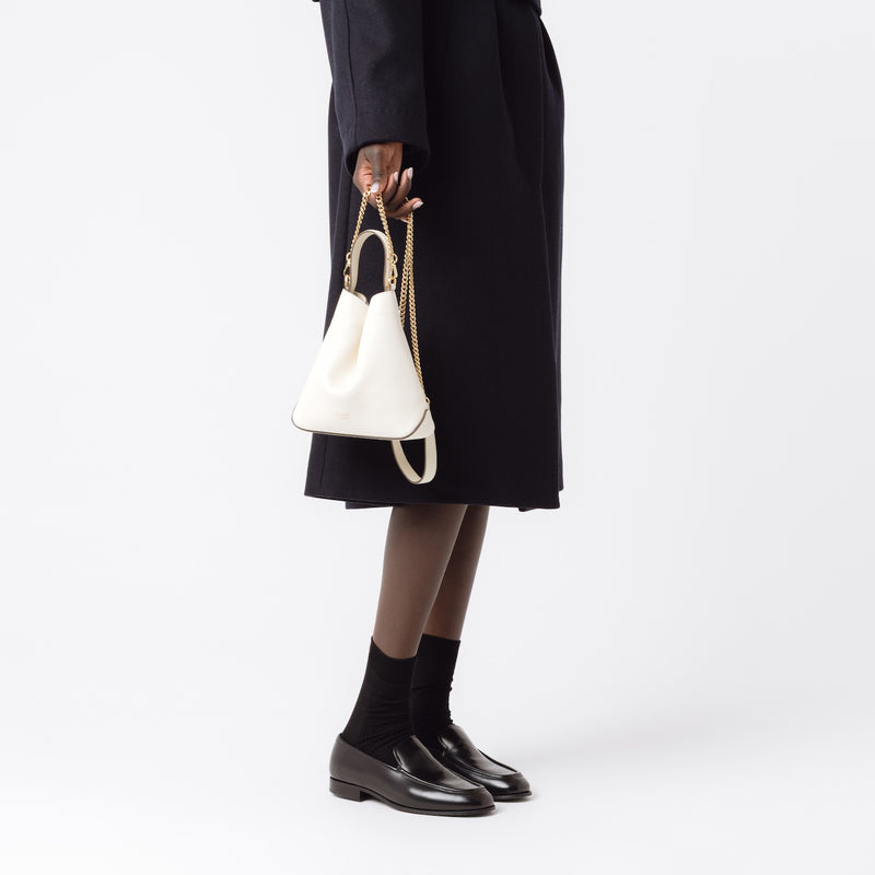 Model holding STOW London's Mini Curve in Spring Moon pebbled leather by the detachable gold chain crossbody strap at her side on a white background 