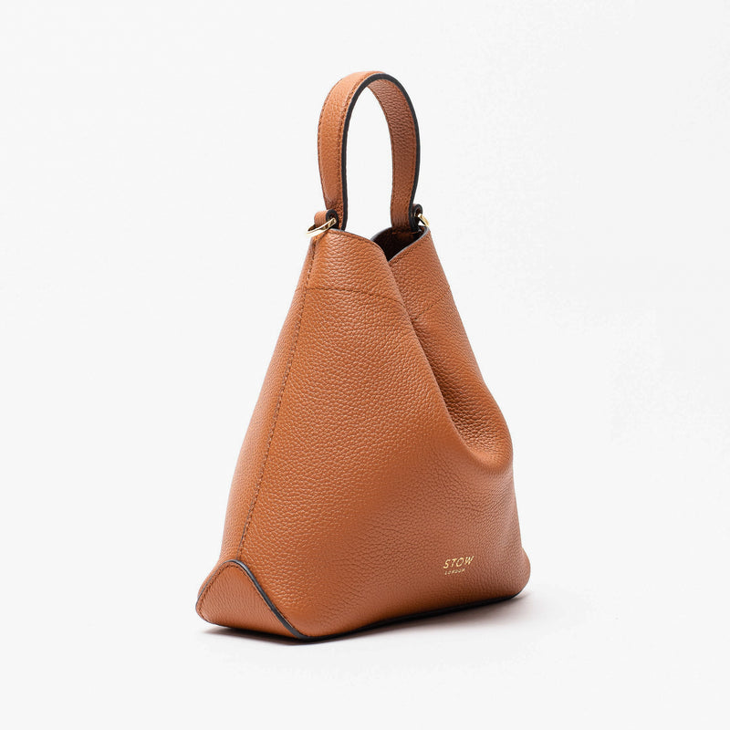 Off-axis view of Stow London's Mini Curve in Earth tan pebbled leather featuring gold details on a light grey background 