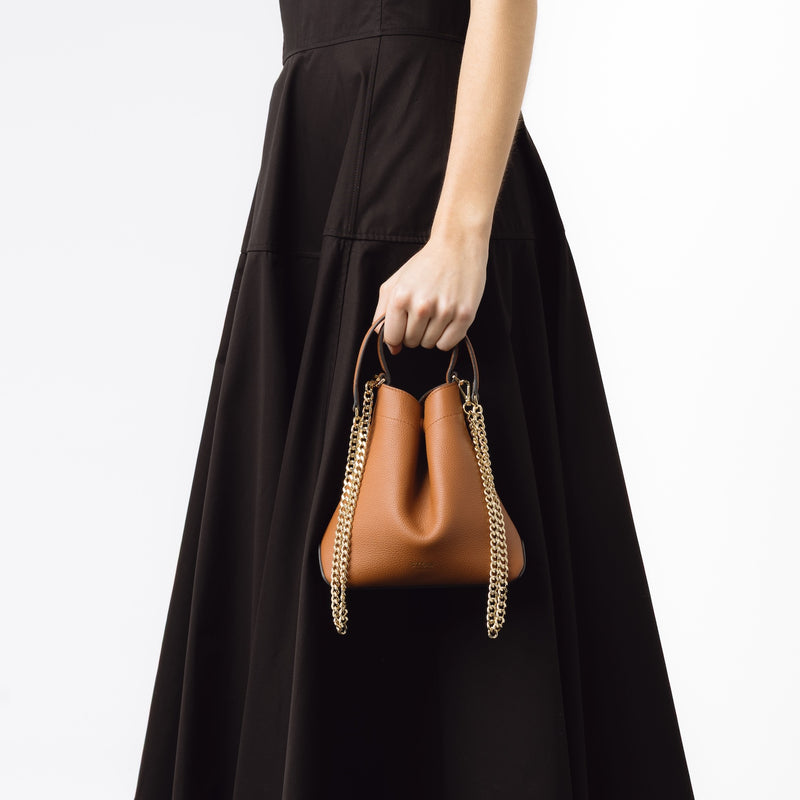 Close shot of model holding STOW London's Mini Curve bag in Earth Tan by its top handle at her side featuring its detachable gold-plated chain on a white background 