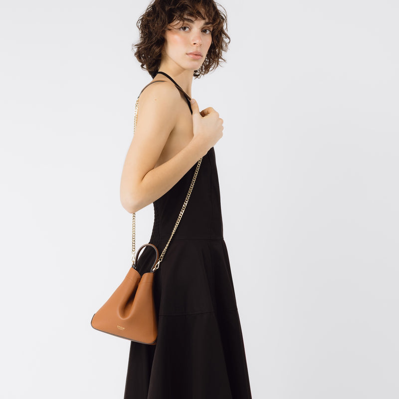 Model wearing STOW London's Mini Curve bag in Earth Tan on her shoulder by its detachable gold-plated chain on a white background 
