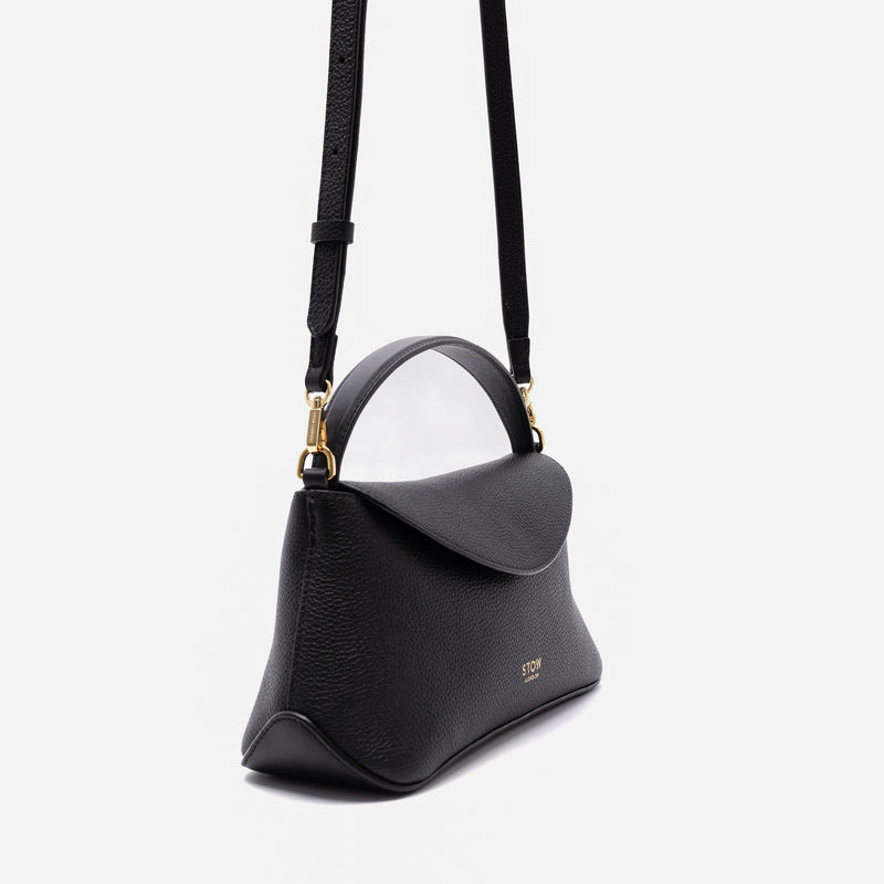 Off-axis view of STOW London's Mini East West in Black pebbled leather and detachable crossbody strap featuring gold-coated stainless steel hardware on a light grey background