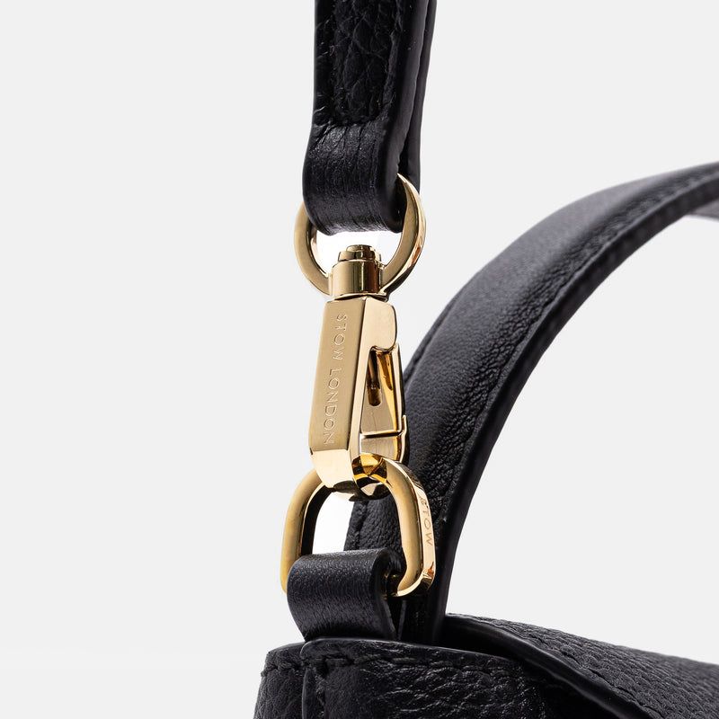Close up of the detachable crossbody strap clip of STOW London's Mini East West in Black pebbled leather featuring gold-coated stainless steel hardware on a light grey background