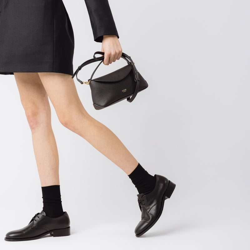 Model holding STOW London's Mini East West in Black pebbled leather featuring gold-coated stainless steel hardware on a light grey background