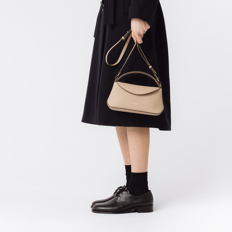 Model holding STOW London's Mini East West in Camel leather by its crossbody strap on a white background