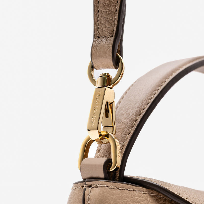 Close up of the detachable crossbody strap clip of STOW London's Mini East West in Camel pebbled leather featuring gold-coated stainless steel hardware on a light grey background