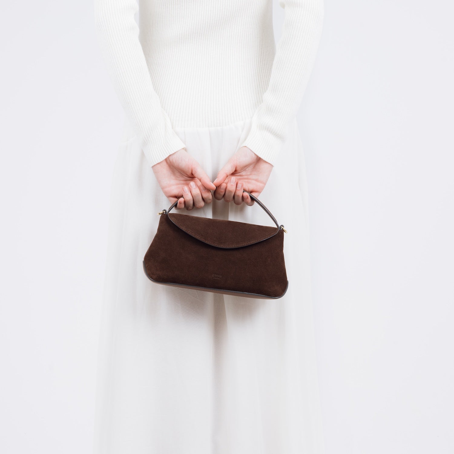 Model holding STOW London's Mini East West bag in Chocolate Suede leather by top handle behind her back featuring gold-coated stainless steel hardware on a white background