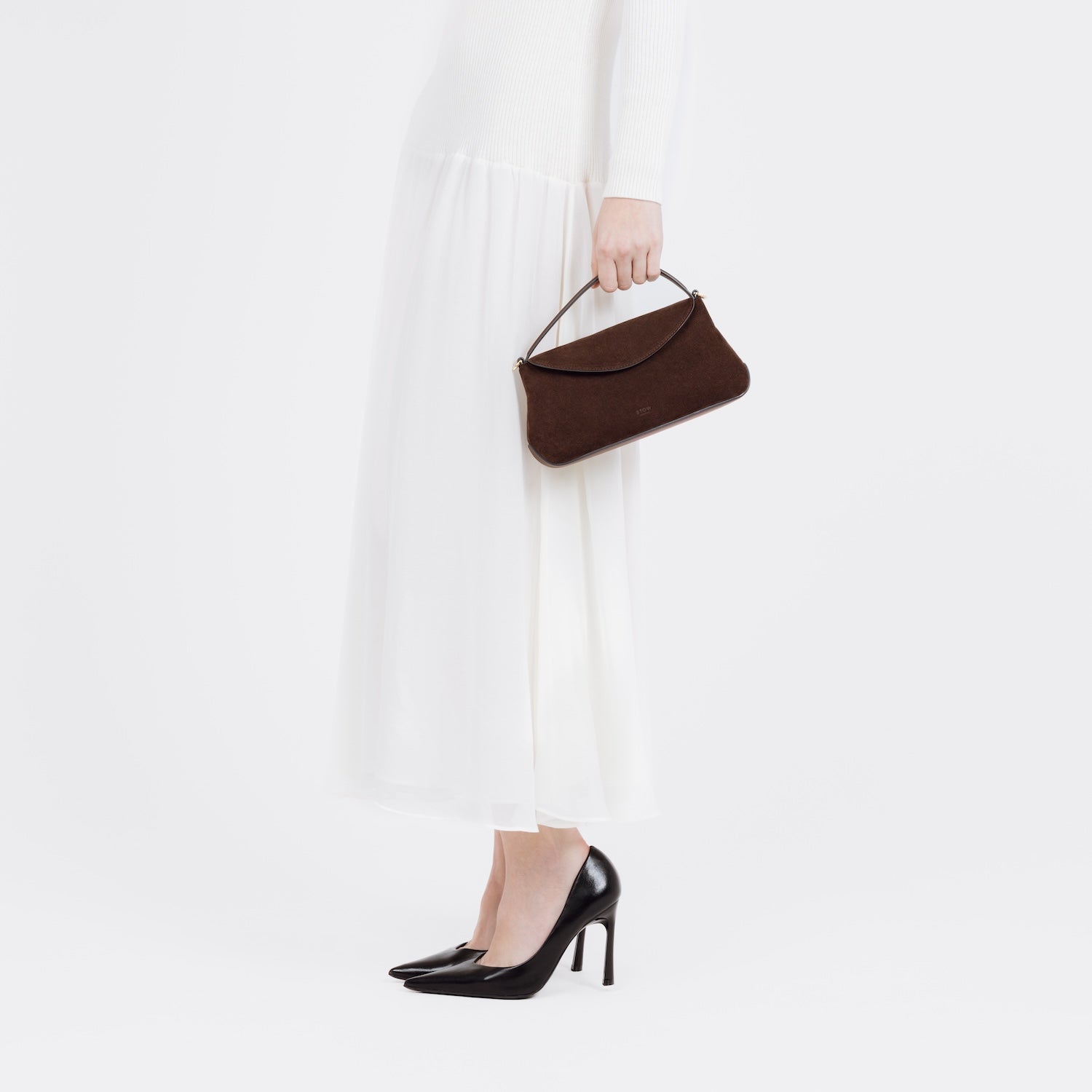 Model holding STOW London's Mini East West bag in Chocolate Suede leather by top handle at her side featuring gold-coated stainless steel hardware on a white background