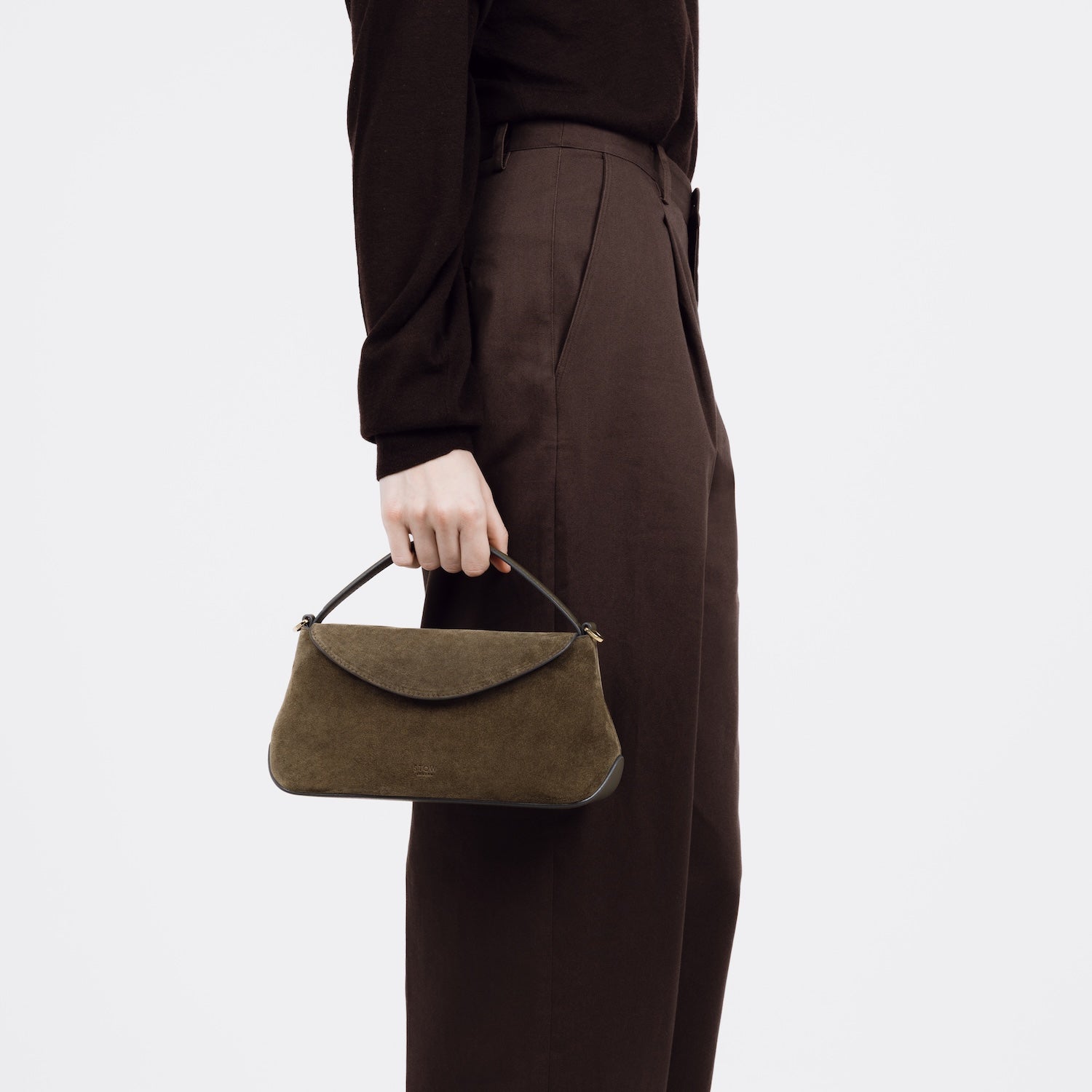 Model holding STOW London's Mini East West bag in Dark Green Suede leather by top handle at her side featuring gold-coated stainless steel hardware on a white background