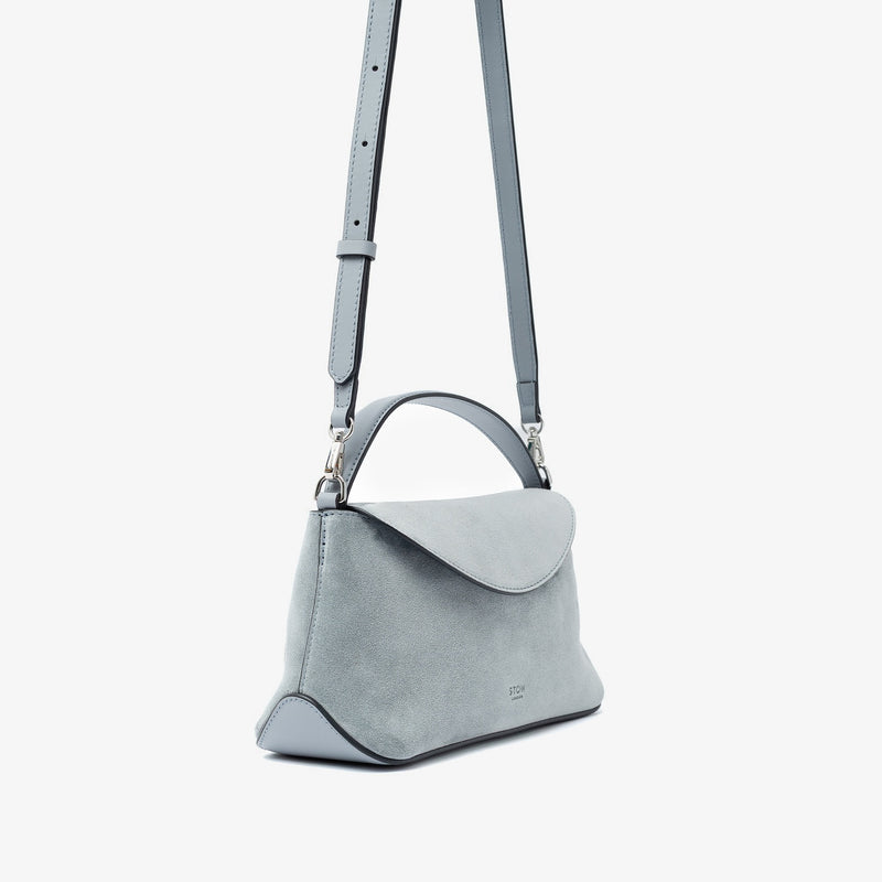 Off-axis view of STOW London's Mini East West in Misty Blue suede leather and detachable crossbody strap featuring silver stainless steel hardware on a light grey background