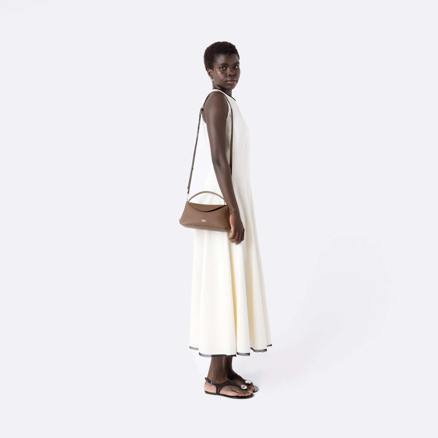 STOW London's Mini East West in Mocha leather worn over shoulder by detachable crossbody strap featuring gold-coated stainless steel hardware on a white background