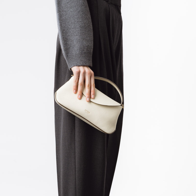 Model holding STOW London's Mini East West in Spring Moon pebbled leather featuring gold-coated stainless steel hardware on a white background