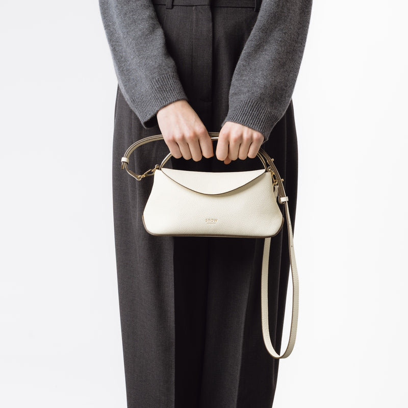 Model holding STOW London's Mini East West in Spring Moon pebbled leather featuring gold-coated stainless steel hardware and detachable crossbody strap on a white background