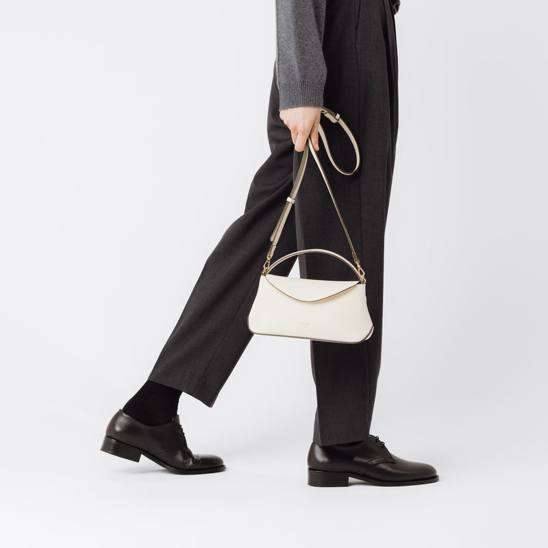 Model holding STOW London's Mini East West in Spring Moon pebbled leather by the detachable crossbody strap featuring gold-coated stainless steel hardware on a white background