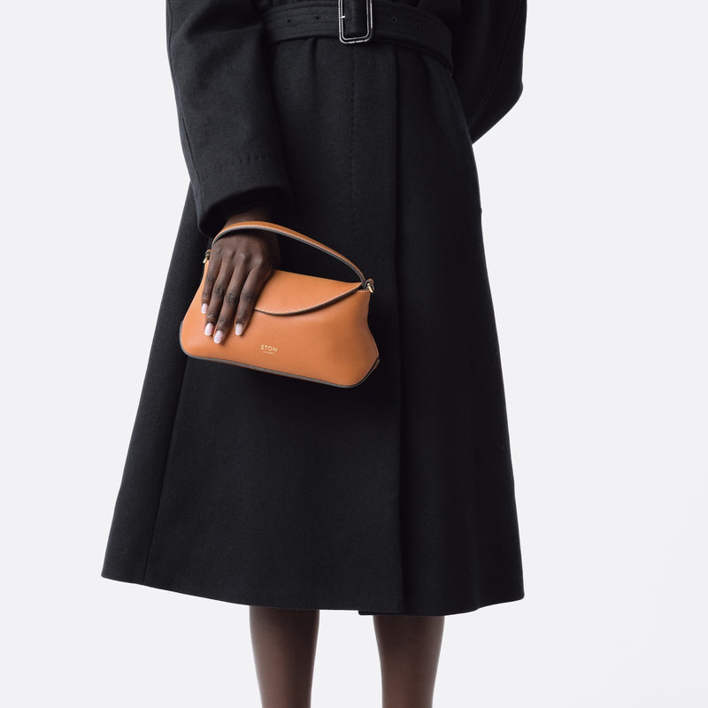 Model holding STOW London's Earth Tan Mini East West bag featuring gold-coated stainless steel hardware on a white background