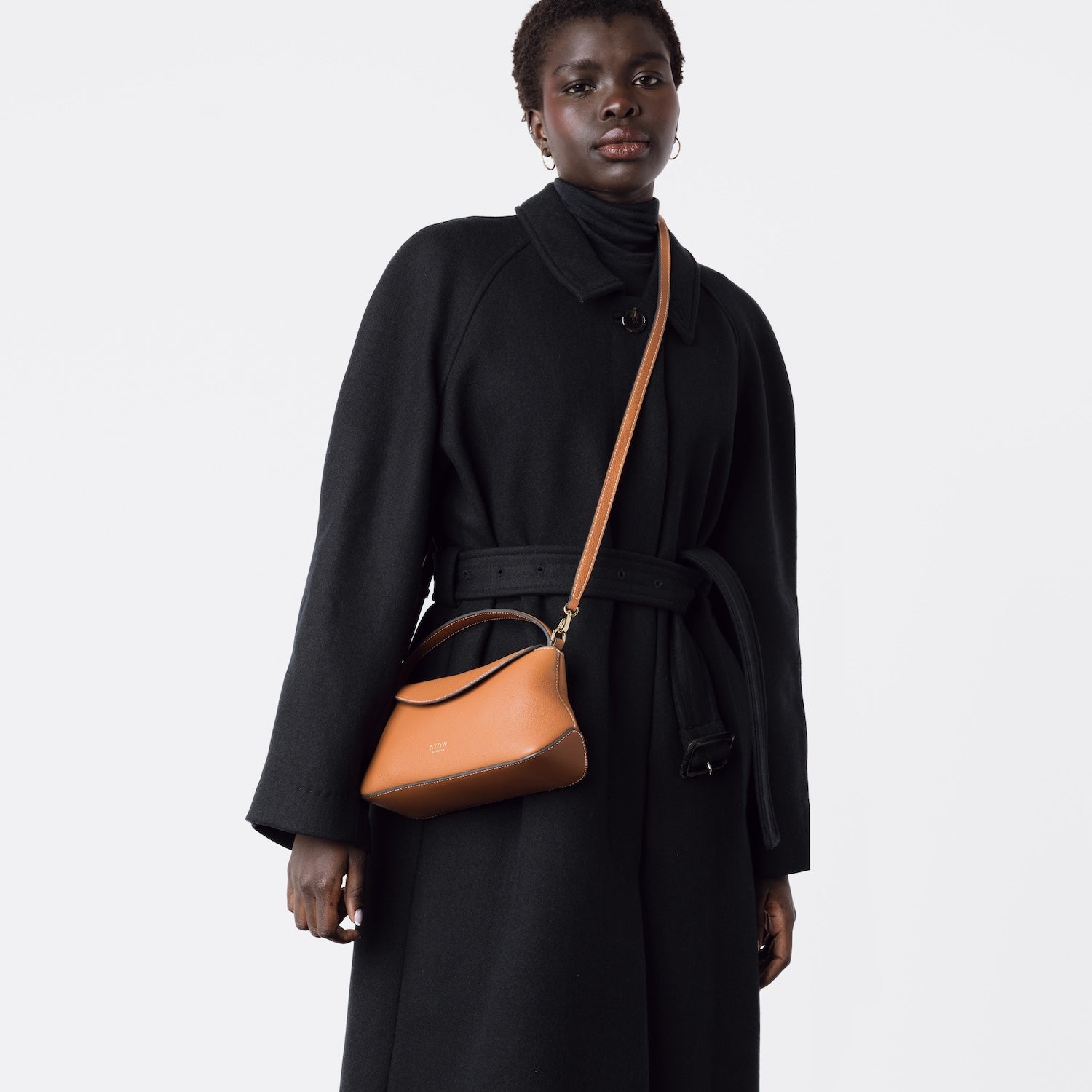 STOW London's Earth Tan Mini East West bag worn crossbody with detachable leather strap by model featuring gold-coated stainless steel hardware on a white background.