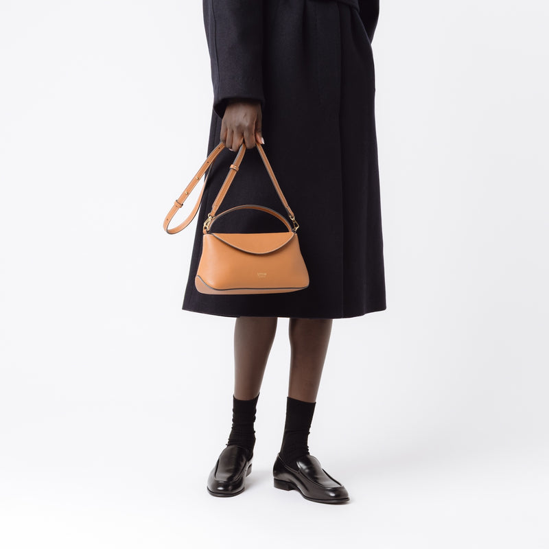 Model holding STOW London's Earth Tan Mini East West bag by detachable leather strap featuring gold-coated stainless steel hardware on a white background.