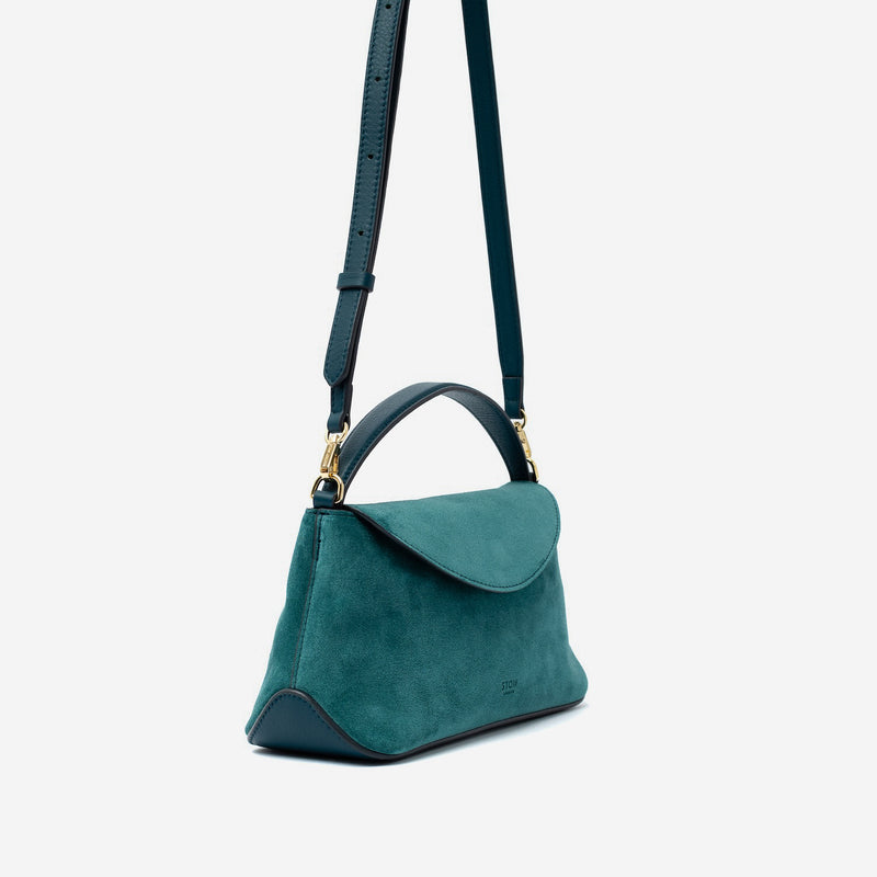 Off-axis view of STOW London's Mini East West in Ocean Teal suede leather and detachable crossbody strap featuring gold-coated stainless steel hardware on a light grey background