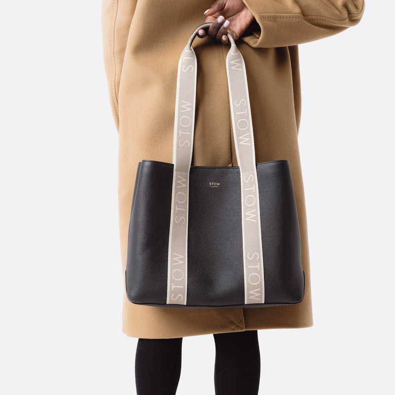 Model holding STOW London's Nastro Tote bag in Black pebbled leather with beige nastro handles featuring 'STOW' branding on a white background