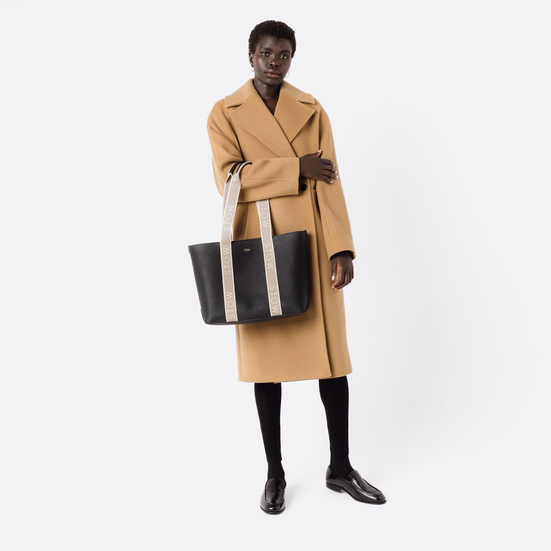 Model holding STOW London's Nastro Tote bag in Black pebbled leather on her arm with beige nastro handles featuring 'STOW' branding on a white background