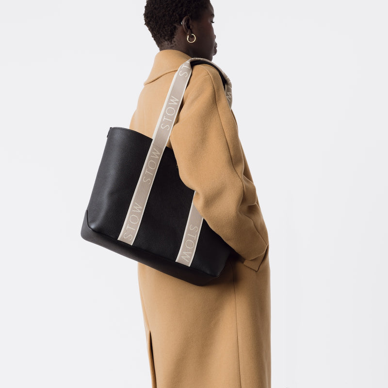 Model wearing STOW London's Nastro Tote bag in Black pebbled leather on her shoulder with beige nastro handles featuring 'STOW' branding on a white background