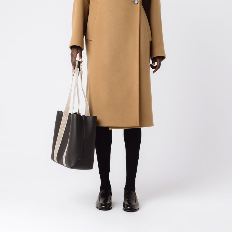 Model holding STOW London's Nastro Tote bag in Black pebbled leather at her side with beige nastro handles featuring 'STOW' branding on a white background