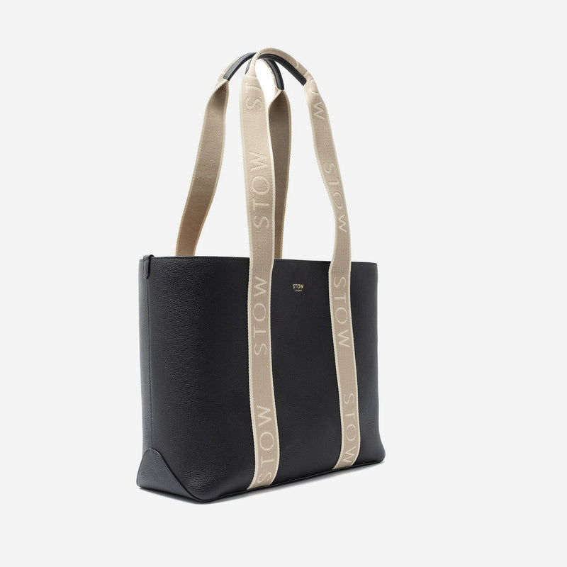 Off-axis view of STOW London's Nastro Tote bag in Black pebbled leather with beige nastro handles featuring 'STOW' branding on a light grey background