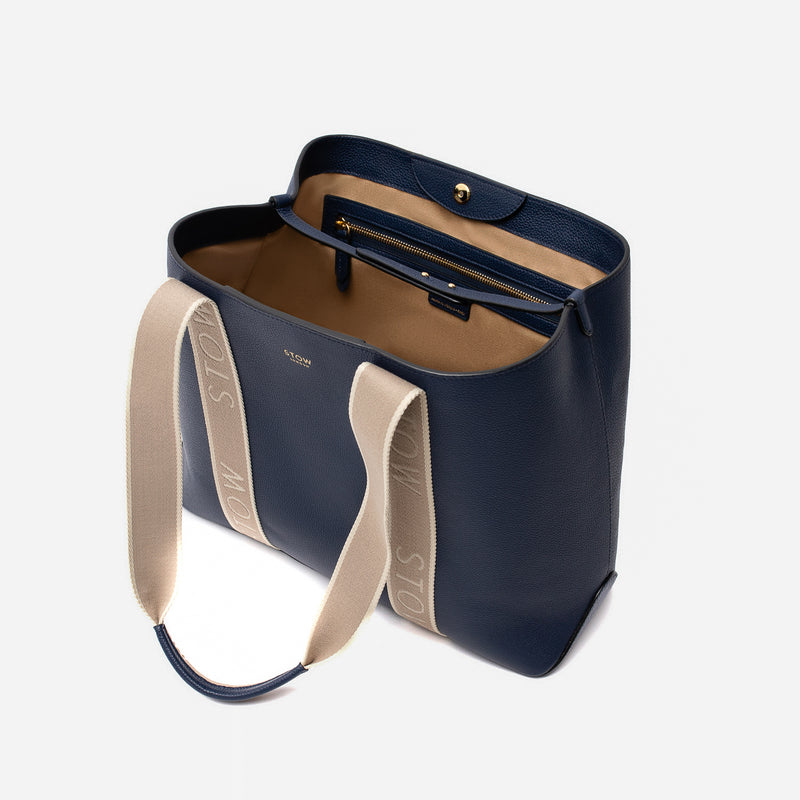 STOW London's Nastro Tote bag in Navy pebbled leather with magnet open showing light beige cotton interior, zip pocket and leather strap on a light grey background